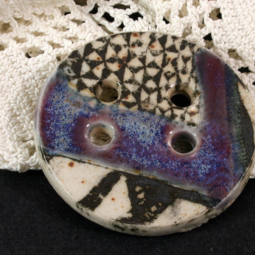 Handmade porcelain button, black graphic against white background, purple/blue accent glaze. (EB198)