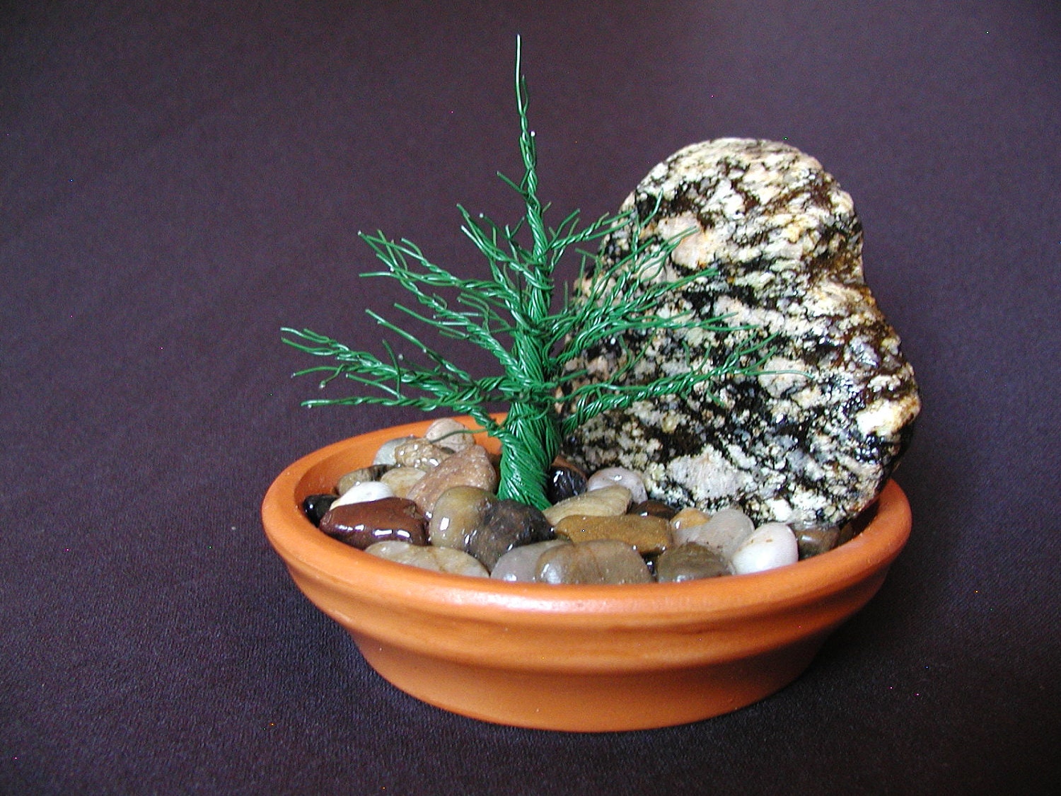 An evergreen grows in river rock near a granite boulder - a mixed media wire tree sculpture.
