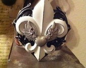 Metal cuff bracelet with aged fleur de lis and accented with a heart with wings